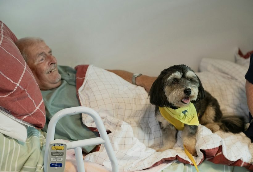 Hombre mayor sonriendo en la cama junto a su perro pequeño, que le hace compañía en casa