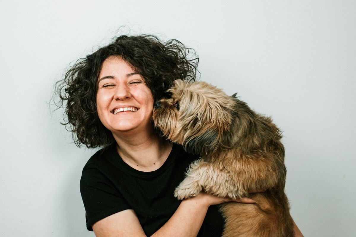Mujer sonriente de cabello rizado abrazando a su perro de apoyo emocional, que le da un beso en la mejilla